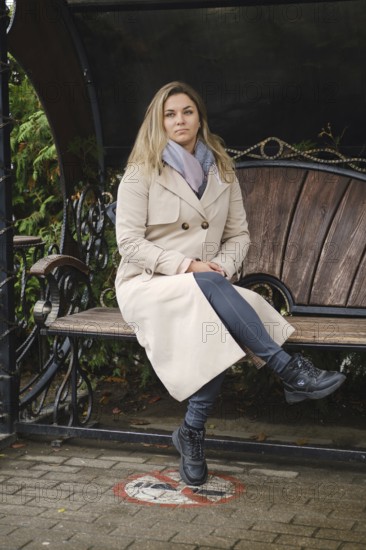 A woman with long hair sits on a bench in a city park. She wears a beige coat, gray scarf, and black boots. Surrounding her are lush green plants, creating a peaceful autumn atmosphere