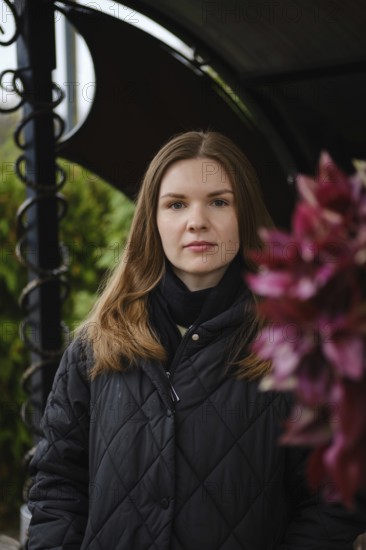 A young woman with long hair poses confidently near vibrant flowers in a garden. The scene captures a serene moment during daylight, surrounded by greenery and natural beauty