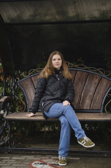 A young woman sits calmly on a wooden bench in a park surrounded by autumn foliage. She wears a black jacket and blue jeans, enjoying the peaceful atmosphere in a serene setting