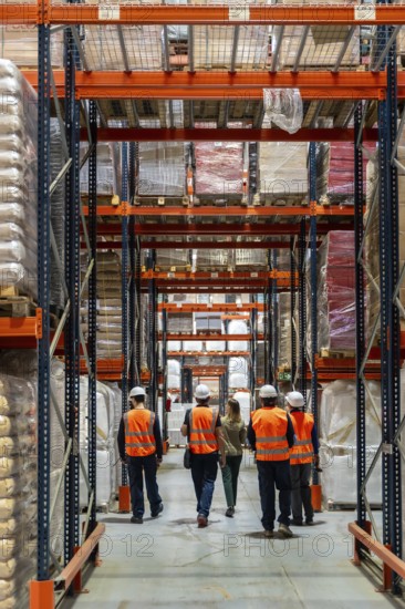 Logistics team walking down a wide warehouse aisle between tall pallet racks, inspecting inventory and coordinating efficient supply chain, safety vests and hardhats visible