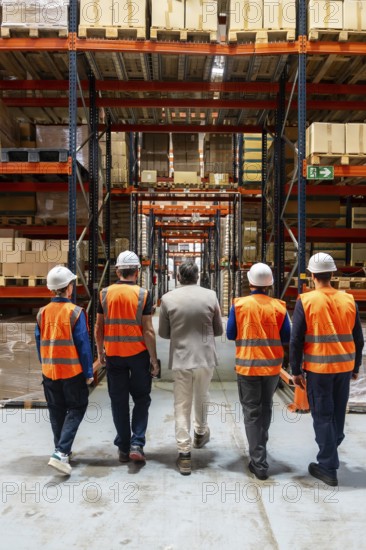 Diverse team of manager and warehouse workers in safety vests and hard hats walking through a logistics facility, inspecting high industrial racks stacked with goods and packages