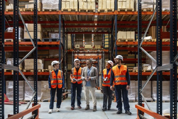 Diverse team of logistics workers and a manager discussing operations and inventory control inside a large industrial warehouse with high shelving racks full of packaged goods