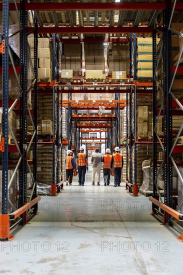 Workers in hard hats and reflective vests walk down a high rack warehouse aisle filled with stacked boxes and pallets, illustrating teamwork, safety and efficient supply chain operations