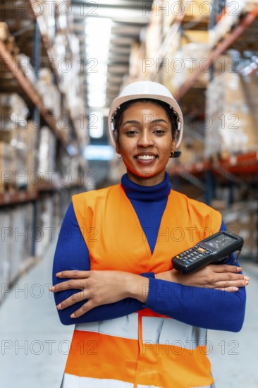 African american woman worker wearing a safety vest and hard hat, holding a portable scanner, standing proudly with arms crossed in a large modern logistics warehouse