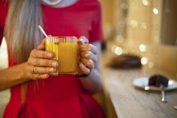 A woman in a bright red dress holds a glass of warm orange drink, enjoying a relaxing moment. Soft lights create a warm atmosphere. In the background, a dessert plate is visible on the table