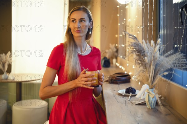 A woman with long hair sits at a bar in a cozy cafe, holding an orange drink. She looks thoughtful as soft lighting and decorative plants create a warm atmosphere around her