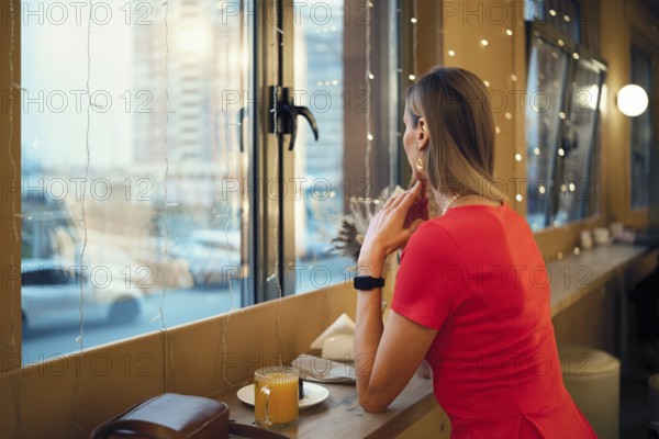 Rear view of a woman dressed in a vibrant red dress sitting near a window in a cozy cafe, looking out at the evening cityscape. A glass of juice rests on the table, reflecting a calm atmosphere