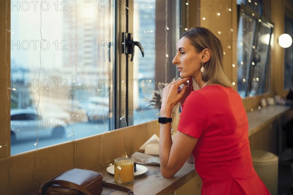 A woman sits quietly in a cafe, gazing out the window at the city skyline. Soft lights surround her as she holds a drink, seeming lost in thought during a peaceful evening