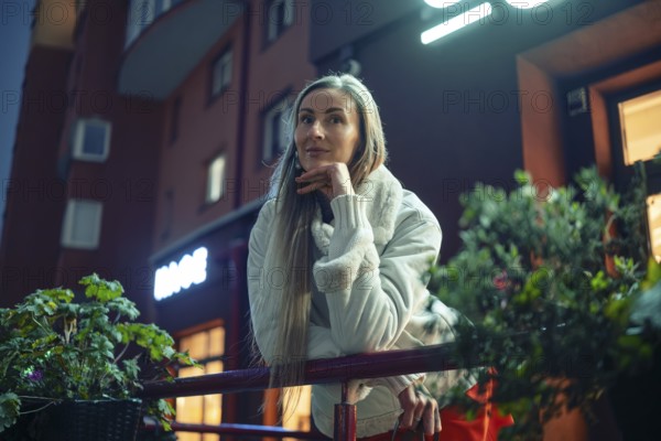 A woman stands thoughtfully on a balcony railing, dressed warmly for the evening chill. The city lights glow softly in the background as she gazes off into the distance, surrounded by greenery