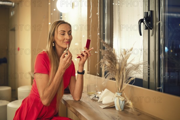 A woman dressed in a bright red dress applies lipstick in front of a mirror. She sits at a wooden counter adorned with soft light, enjoying an evening in a stylish space filled with plants