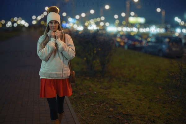 A woman walks on a city sidewalk at night, wearing a warm white jacket and a beanie. The street is filled with bright lights and parked cars, creating a lively urban atmosphere
