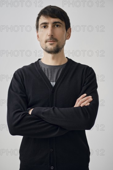 A man stands with arms crossed, portraying confidence. He wears a black cardigan over a gray shirt. The background is simple and light, emphasizing his serious expression during the day
