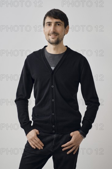 A man stands confidently with his hands on his hips, wearing a black cardigan over a t-shirt. The background is light, creating a bright and welcoming atmosphere