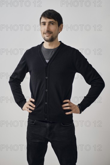 A man poses confidently with his hands on his hips, wearing a black cardigan over a gray shirt. He has short hair and stands in a simple studio with a plain background