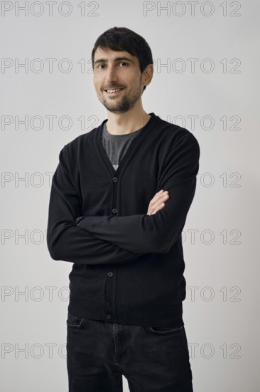 A man is posing confidently with arms crossed, wearing a black cardigan over a gray shirt. He stands against a simple, light backdrop, giving a relaxed yet professional appearance