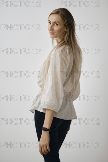 A woman turns back showcasing her long hair and stylish outfit. She gazes over her shoulder, exuding elegance and poise in a simple indoor setting with a neutral backdrop