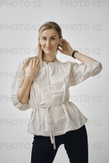 A playful woman stands in a bright studio, wearing a fashionable light blouse and dark pants. She smiles while playing with her hair, showcasing a relaxed and cheerful mood