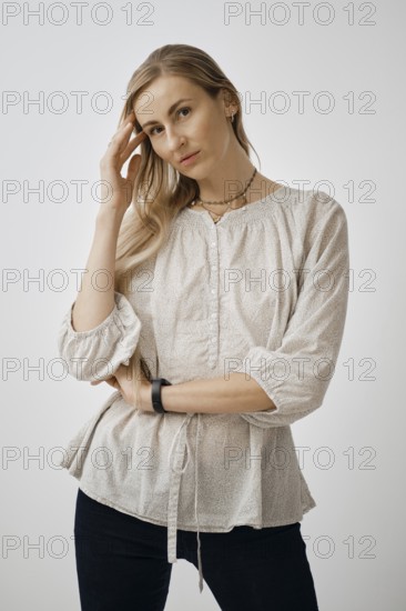 A pensive woman is standing with one hand touching her hair while wearing a light, textured blouse. She is indoors in a simple background, capturing a casual look