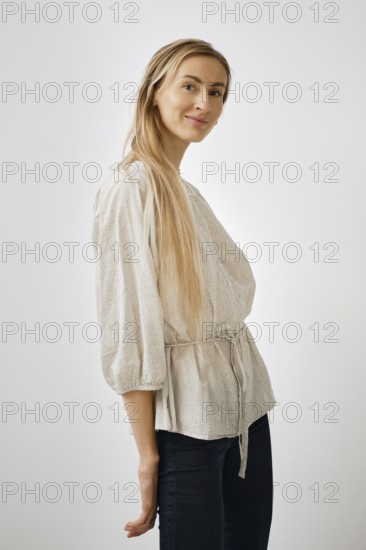 A blonde woman model stands playfully in a light-colored blouse with bell sleeves. She wears dark jeans and poses against a simple backdrop, showcasing her outfit and hairstyle
