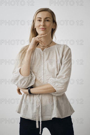 A woman stands in a relaxed pose with one hand on her chin, wearing a light-colored, casual blouse. She looks relaxed with a slight smile, set against a simple, neutral background