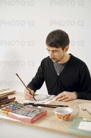 A male artist focuses on his work at a wooden table filled with various art supplies. He uses paintbrushes and colorful materials to create an intricate design in a well-lit, simple space
