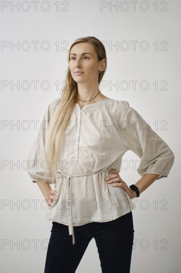 A woman stands confidently in a light beige blouse with long sleeves, paired with dark jeans. Her hair is styled long and straight, and she poses gracefully, showcasing modern fashion