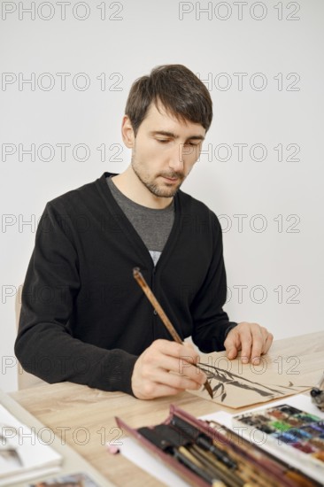 A man carefully draws on a sheet of paper in a bright studio. He uses a brush and appears deep in thought. Art supplies are scattered around him, enhancing the creative atmosphere
