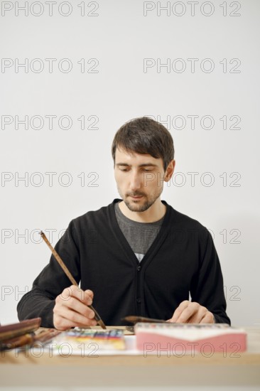 A focused artist is working diligently on a paper in a clean, bright studio. He uses a brush to add detail to his artwork surrounded by paints and brushes