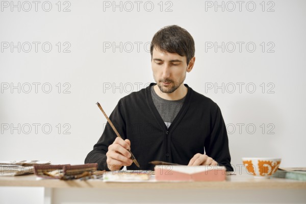 A man is concentrating on painting in a minimalist studio, surrounded by art supplies and colorful materials. Natural light fills the room, enhancing the creative atmosphere of his work