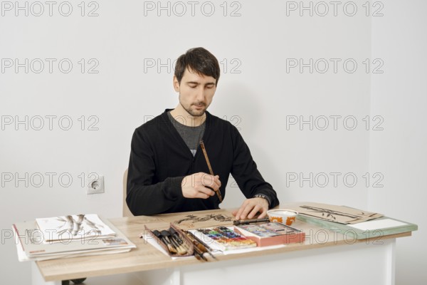 An artist sits at a wooden table, focused on painting with watercolors. The studio is bright and minimalistic, filled with art supplies and sketches. The atmosphere is calm and creative
