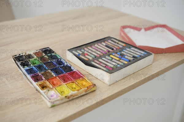 A wooden table displays two sets of art supplies. On the left, a vibrant watercolor palette with many colors rests beside a box of pastels. Perfect for artists and creative enthusiasts