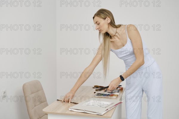 A woman dressed in light clothing is focused on arranging her art supplies and sketches on a table in a well-lit, minimalistic room. She is preparing for a creative session
