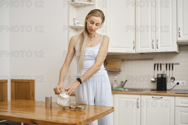 A woman in light clothing pours tea from a kettle into a bowl on a wooden table. The bright kitchen has wooden accents and is simple yet inviting, perfect for a calm afternoon tea time