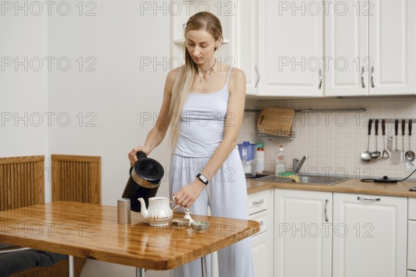A woman stands in a modern kitchen pouring hot water from a kettle into a teapot. She wears a light outfit and is preparing to brew tea, focused on her task amidst the cozy surroundings