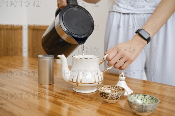 A person pours hot water from a kettle into a delicate teapot. On the wooden table, small bowls are placed, displaying traditional designs