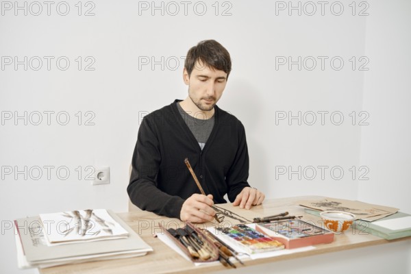 A focused artist is drawing at a wooden table in a bright studio. Various art supplies, including brushes and paints, are neatly arranged nearby, creating an inspiring workspace for creativity
