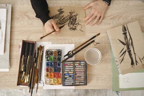 A person is painting with watercolors and sketching with fine brushes on brown paper. Art supplies are organized on a wooden table, including paints, brushes, and a coffee cup nearby