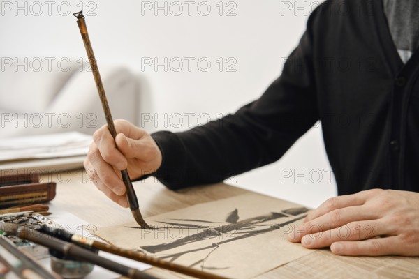 An artist is focused on creating a detailed brush painting on paper. The scene shows natural light streaming in, highlighting the tools and materials used for this traditional art form