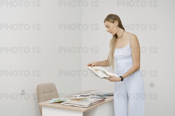 A woman with long hair stands in a bright, minimal room, looking through art sketches. She focuses on the details of the drawings while surrounded by various creative works on the table