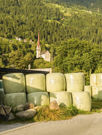 Agricultural structures with hay bales sealed in plastic near a farm on the outskirts of Oetz in Ötztal, Tyrol, Austria, with the parish church in the background