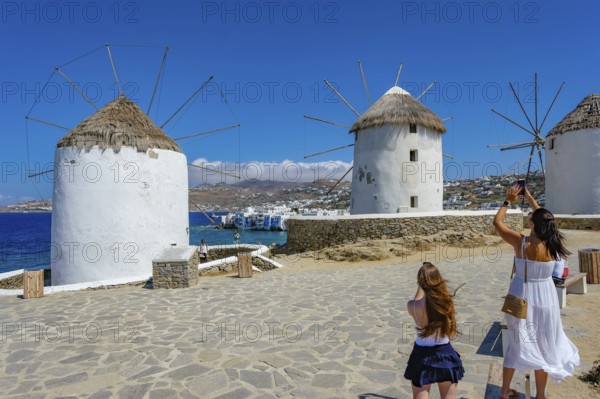 Mykonos, Cyclades, Greece - Tourists photograph the six sixteenth-century windmills lined up on a hill above Mykonos Town, Mykonos Chora. The windmills are the island's landmark. At the back, right by the sea, are the colorful houses of Little Venice. Mykonos is part of the Cyclades archipelago in the Aegean Sea