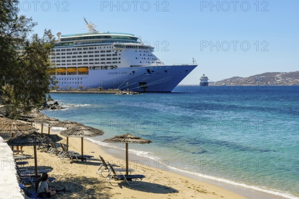 Mykonos, Cyclades, Greece - Voyager of the Seas cruise ship is located in Mykonos Harbour Bay. Front beach chairs and umbrellas on the sandy beach of Paralia Agios Stefanos