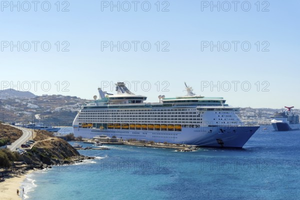 Mykonos, Cyclades, Greece - Voyager of the Seas cruise ship is located in Mykonos Harbour Bay. In front, tourists bathe on the sandy beach of Paralia Agios Stefanos. In the back is the old town of Mykonos