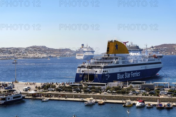 Mykonos, Cyclades, Greece - Mykonos New Port, a Blue Star Ferries ferry, is located on the quay in the new port in front of the skyline of Mykonos Town. In front, a taxi boat a sea bus takes tourists to the old town. At the back, there are more cruise ships in the bay in front of Mykonos City with a view of the old town with the windmills. Cruise ships bring tourists to the old town