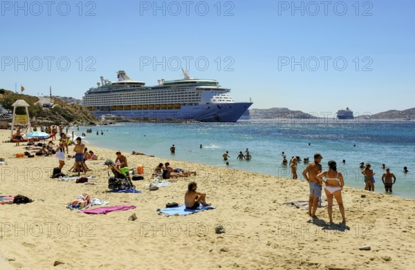 Mykonos, Cyclades, Greece - Voyager of the Seas cruise ship is located in Mykonos Harbour Bay. In front, tourists and locals bathe on the sandy beach of Paralia Agios Stefanos