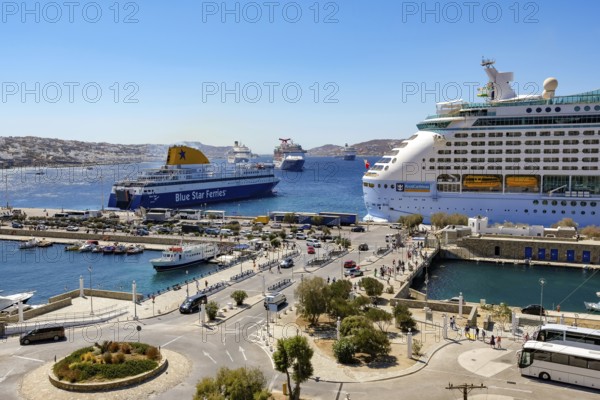 Mykonos, Cyclades, Greece - Mykonos New Port, a Blue Star Ferries ferry and a cruise ship are located on the quay in the new port in front of the skyline of Mykonos Town. At the back, there are more cruise ships in the bay off Mykonos City with a view of the old town with the windmills. Cruise ships bring tourists to the old town