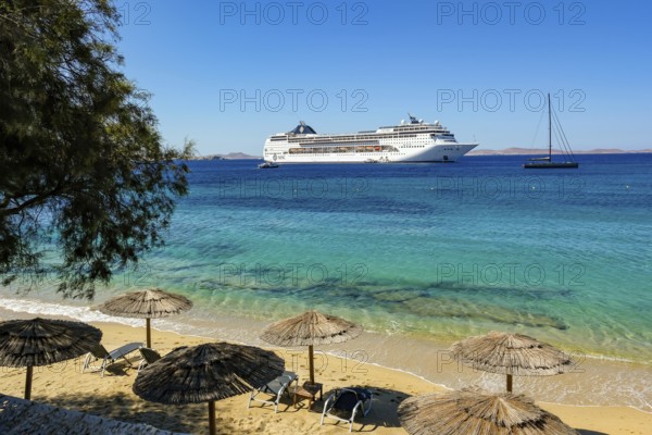 Mykonos, Cyclades, Greece - Cruise ship MSC Lirica is located in Mykonos Harbour Bay. Cruise ships bring tourists to shore. In front, deserted beach chairs and umbrellas on the sandy beach of Paralia Agios Stefanos