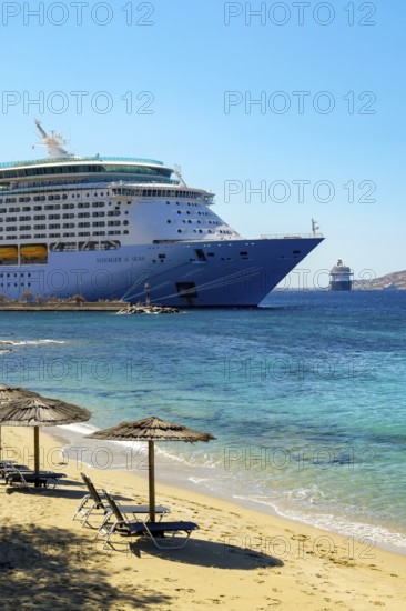 Mykonos, Cyclades, Greece - Voyager of the Seas cruise ship is located in Mykonos Harbour Bay. Front beach chairs and umbrellas on the sandy beach of Paralia Agios Stefanos