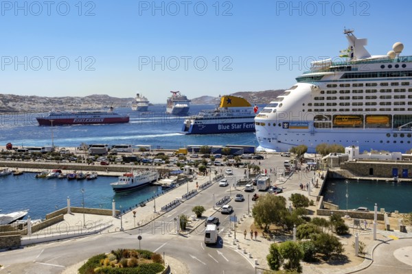 Mykonos, Cyclades, Greece - Mykonos New Port, Blue Star Ferries and Fast Ferries and a cruise ship are moored on the quay in the new port in front of the skyline of Mykonos Town. At the back, there are more cruise ships in the bay off Mykonos City with a view of the old town with the windmills. Cruise ships bring tourists to the old town