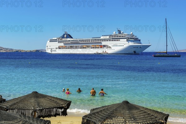 Mykonos, Cyclades, Greece - Cruise ship MSC Lirica is located in Mykonos Harbour Bay. Cruise ships bring tourists to shore. In front, tourists bathe on the sandy beach of Paralia Agios Stefanos